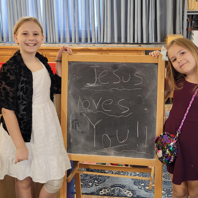 Two children with chalkboard inscribed with "Jesus Loves You"
