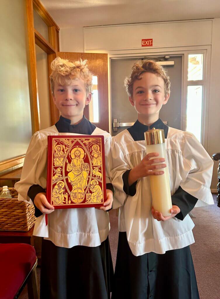 Two young acolytes in white surplices over black cassocks stand inside a church smiling at the camera. One holds an ornate red Gospel book with gold iconography of Christ, while the other holds a tall white candle with a gold top used in the service. An open doorway and exit sign are visible behind them.