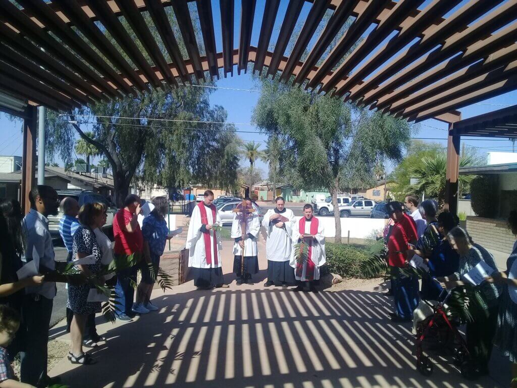 Clergy in white vestments with red stoles lead a Palm Sunday procession outdoors beneath a wooden pergola while congregants stand on both sides holding palm branches and service leaflets. Bright sunlight casts striped shadows across the walkway, and neighborhood homes and trees are visible in the background.